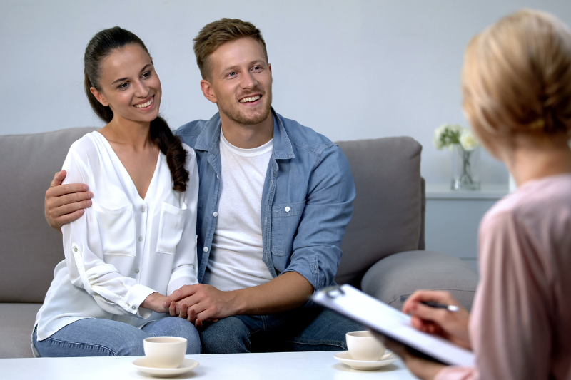 A smiling couple sits close together on a couch, holding hands and talking to a person with a clipboard, possibly a therapist or counselor, in a comfortable room with coffee cups on a table.