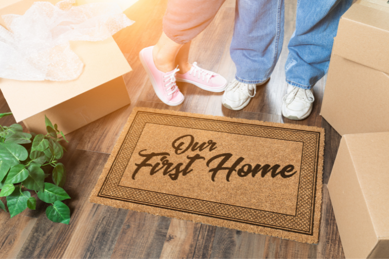 Two people stand next to a doormat that reads Our First Home, surrounded by moving boxes and a plant on a wooden floor, with sunlight shining in.