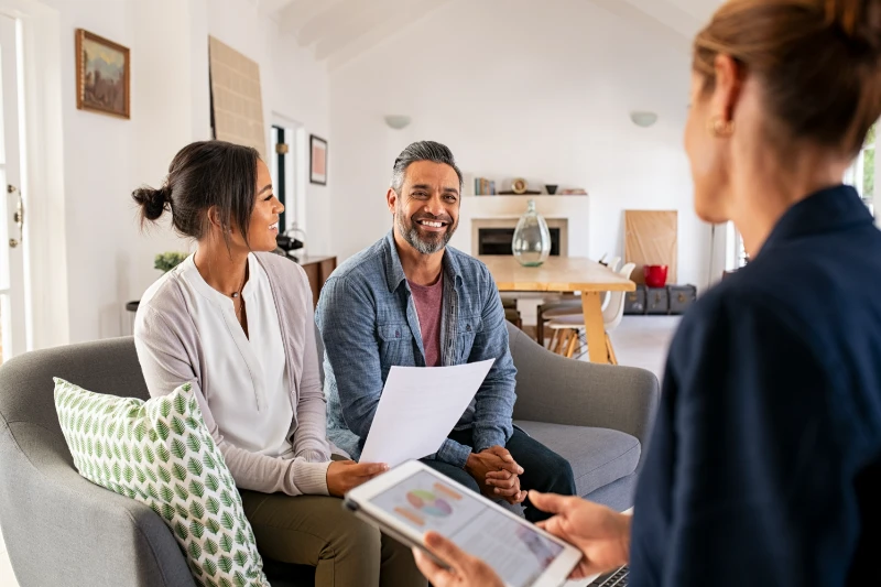 Reverse Mortgage 1 A smiling couple sits on a couch holding documents while talking to a professional woman with a digital tablet in a bright, modern living room.